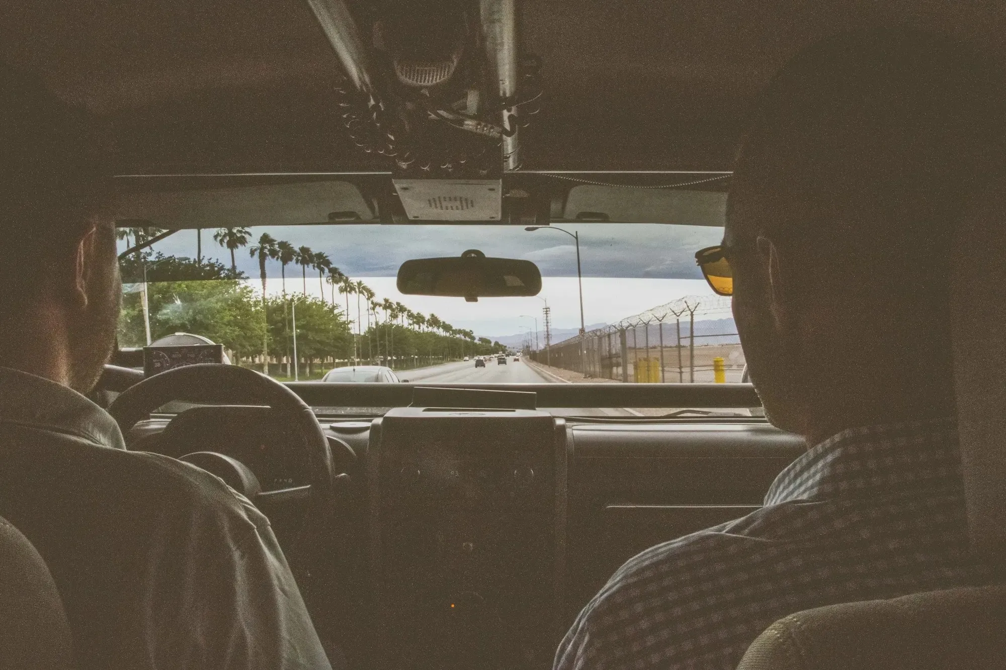 The view through the windscreen of a car from the back seat, two people in the front, a palm-lined road ahead.